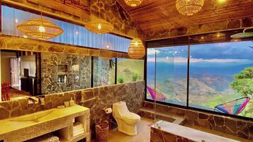 A bathroom with a stone wall, a toilet, a sink, and a large window with a view of the landscape.
