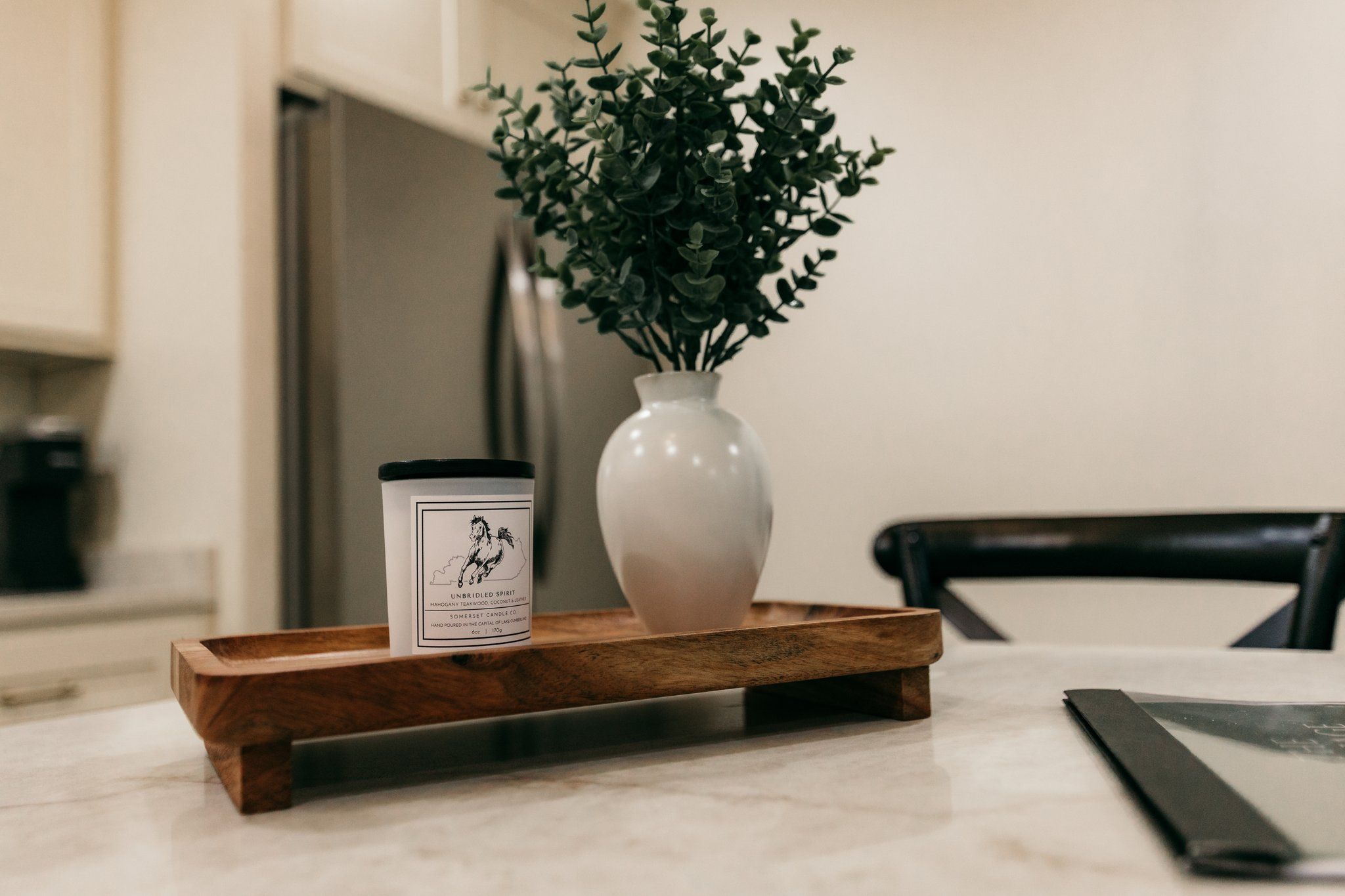 Un vase blanc orné de feuillage vert, posé sur un plateau en bois, une bougie décorée d’une illustration de cheval et une chaise noire, le tout dans un décor de cuisine.
