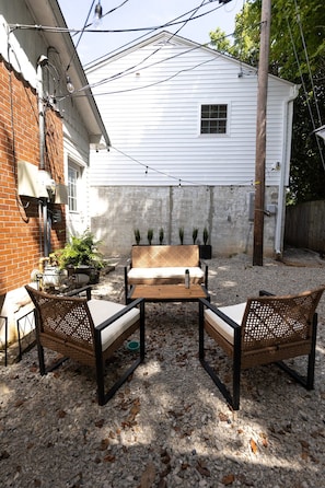 An outdoor patio with a wooden bench and two wicker chairs, a potted plant, and a small table.