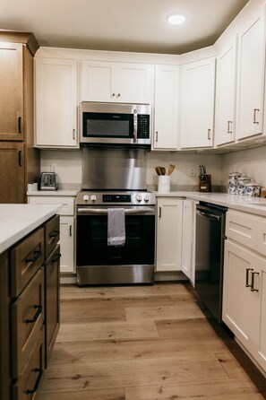 A kitchen with white cabinets, a stainless steel microwave, an oven, a dishwasher, and wooden flooring.