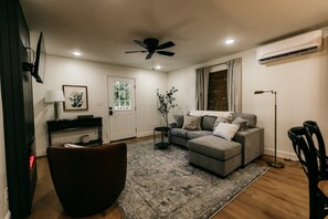 A living room with a grey sofa, a brown armchair, a black floor lamp, a ceiling fan, and a window with curtains.