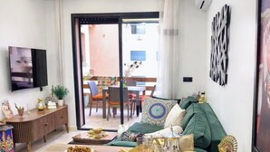 Ein modernes Wohnzimmer mit einer zentralen Kücheninsel, zwei Barhocker, einem Flachbildfernseher und einem Essbereich, der durch eine Glastür sichtbar ist.