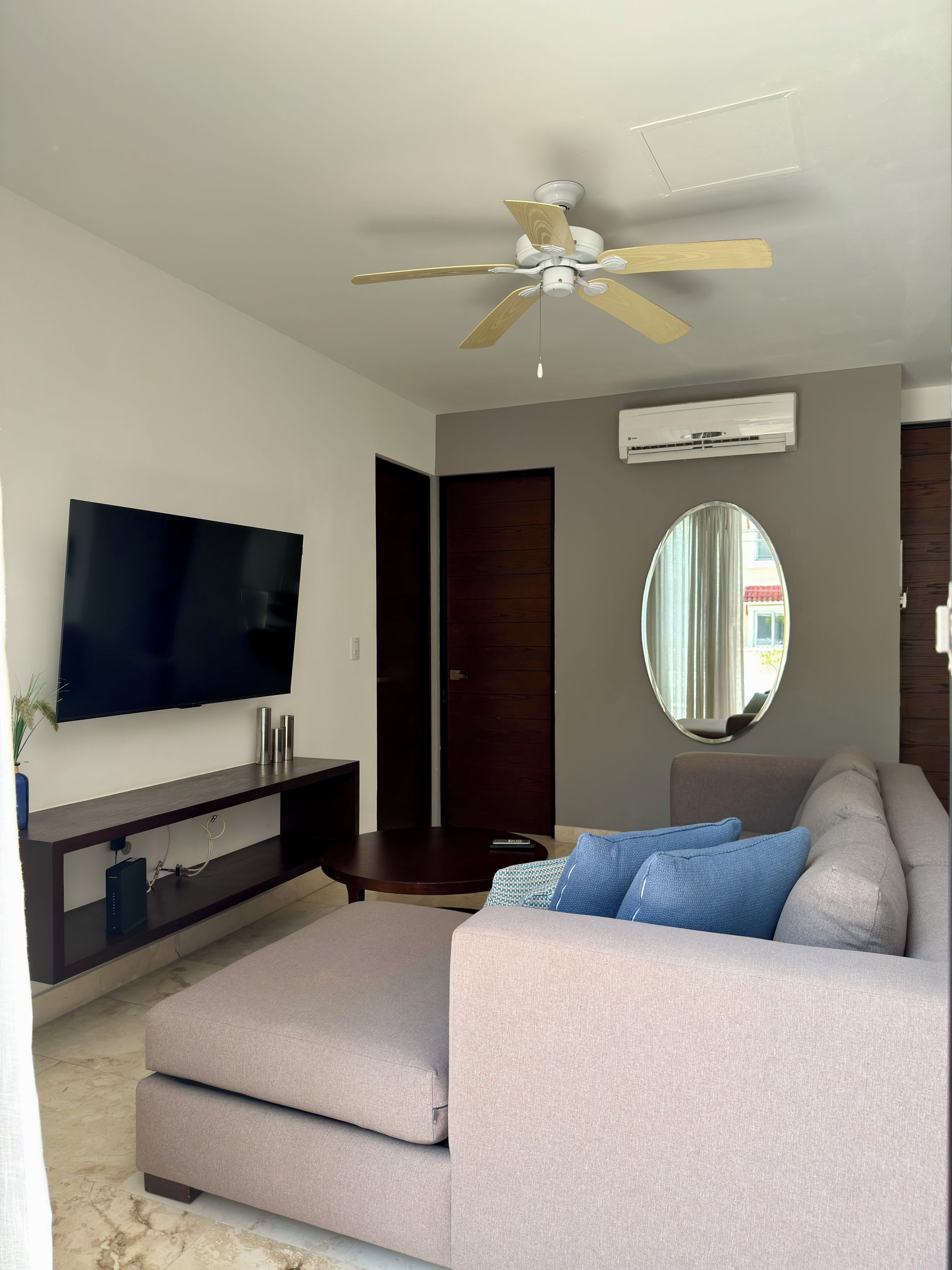 Ein modernes Wohnzimmer mit Flachbildfernseher, Deckenventilator, rundem Spiegel und einer blauen Sofabezüge.