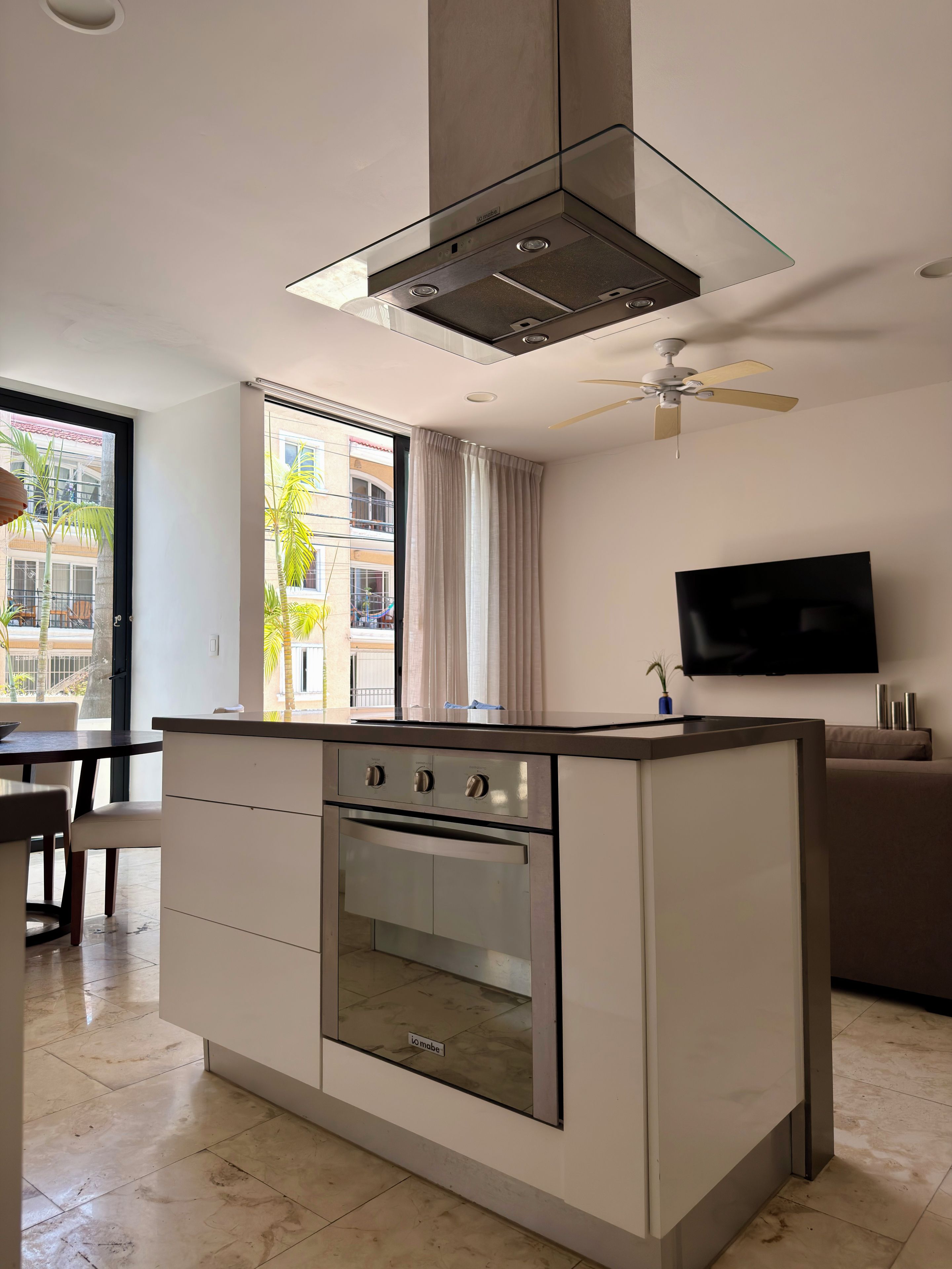 A modern kitchen with a built-in oven, a dining area with a table and chairs, a large window with curtains, and a ceiling fan.