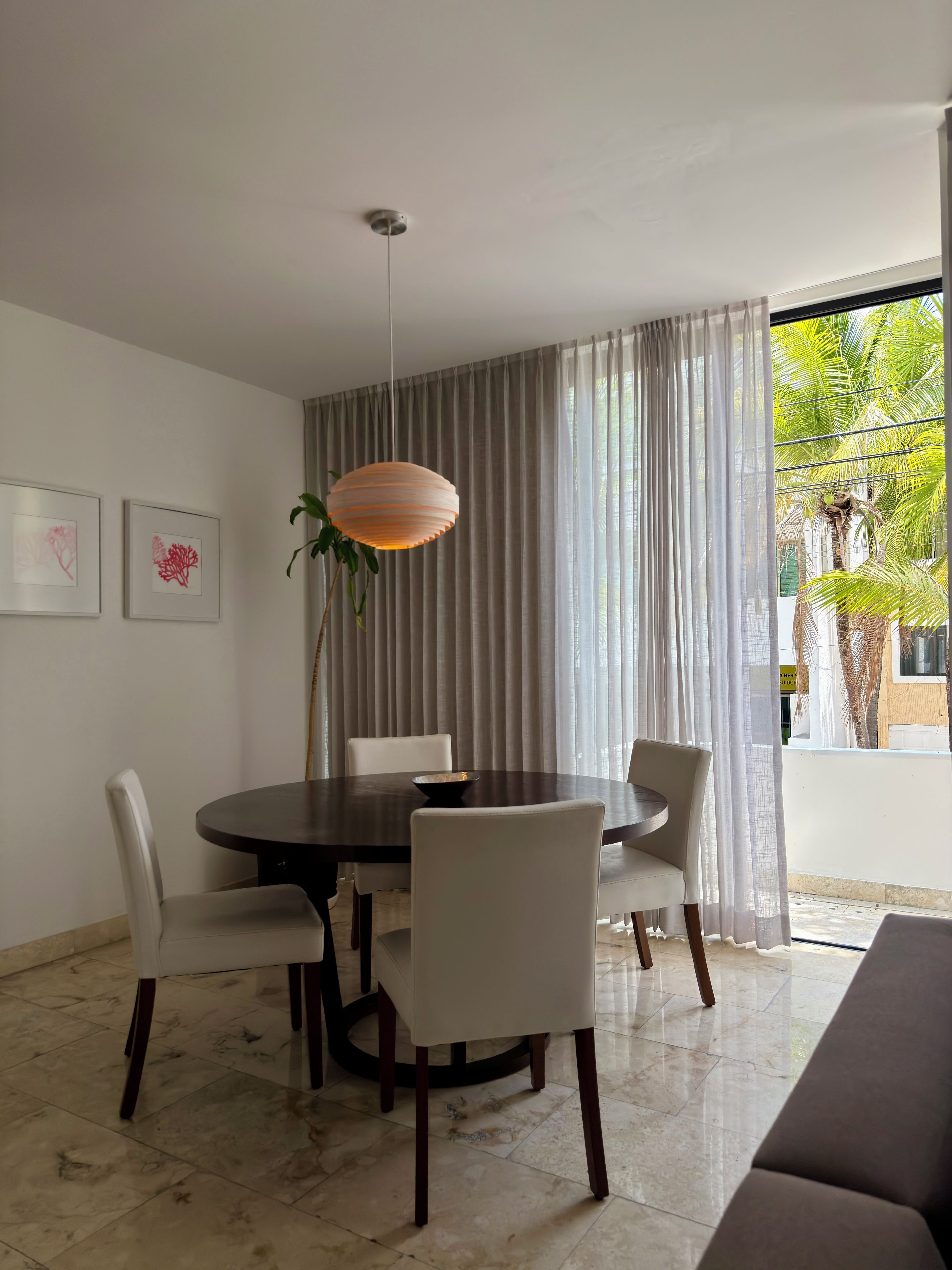 A dining area with a round table, four chairs, a hanging light fixture, and a view of palm trees through sheer curtains.