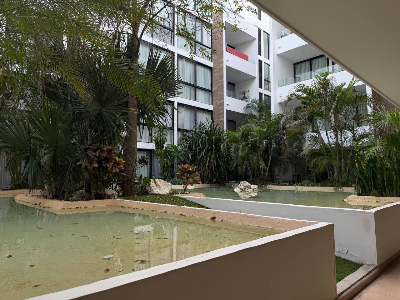 A modern building with multiple balconies, surrounded by lush tropical plants and a small pool area.