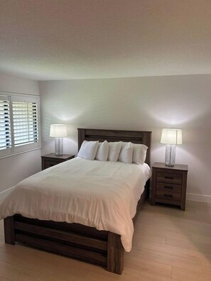 Un dormitorio con una cama de madera, dos mesitas de noche con lámparas, una ventana con persianas y un piso de color claro.