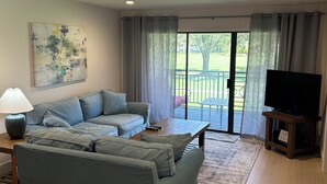 A living room with a sectional sofa, a coffee table, a TV on a stand, a painting on the wall, and a sliding glass door leading to an outdoor area. - Cozy 2-bed/2 bath condo in Saddlebrook Resort (Zephyrhills)