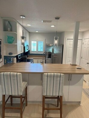 Una cocina con una isla central, dos taburetes de bar, iluminación colgante y vista al comedor.