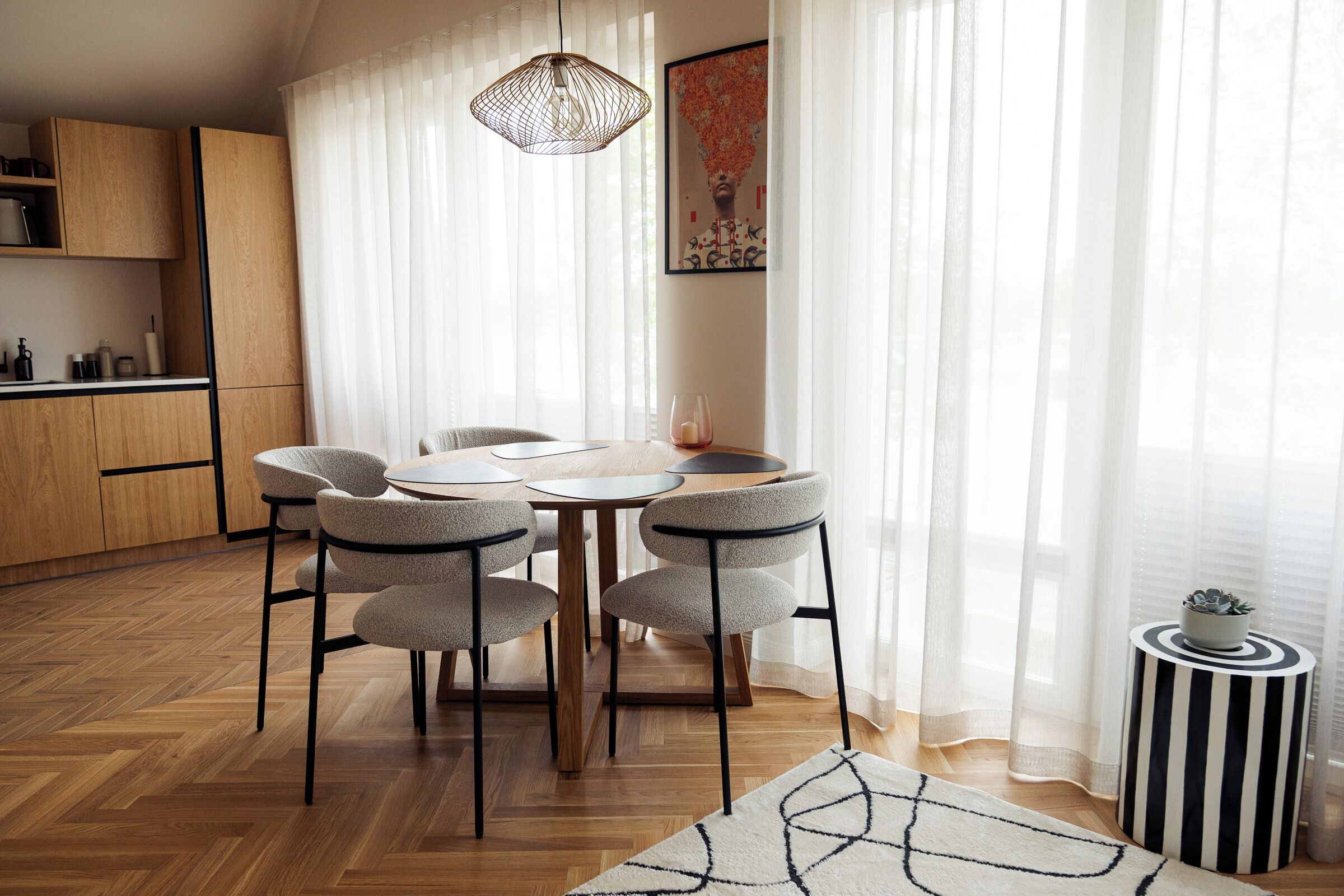 A dining area with a round table, four chairs, a hanging light fixture, a kitchen area with wooden cabinets, and a small side table with a plant.