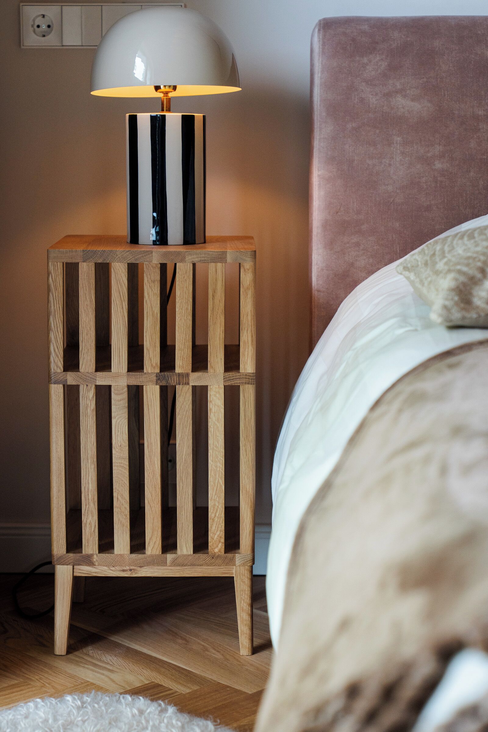 A wooden nightstand with a lamp, a bed with a headboard, and a white rug.