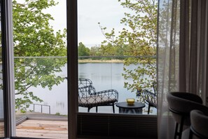 A view from a room with a large window overlooking a lake, featuring outdoor furniture and a small table.