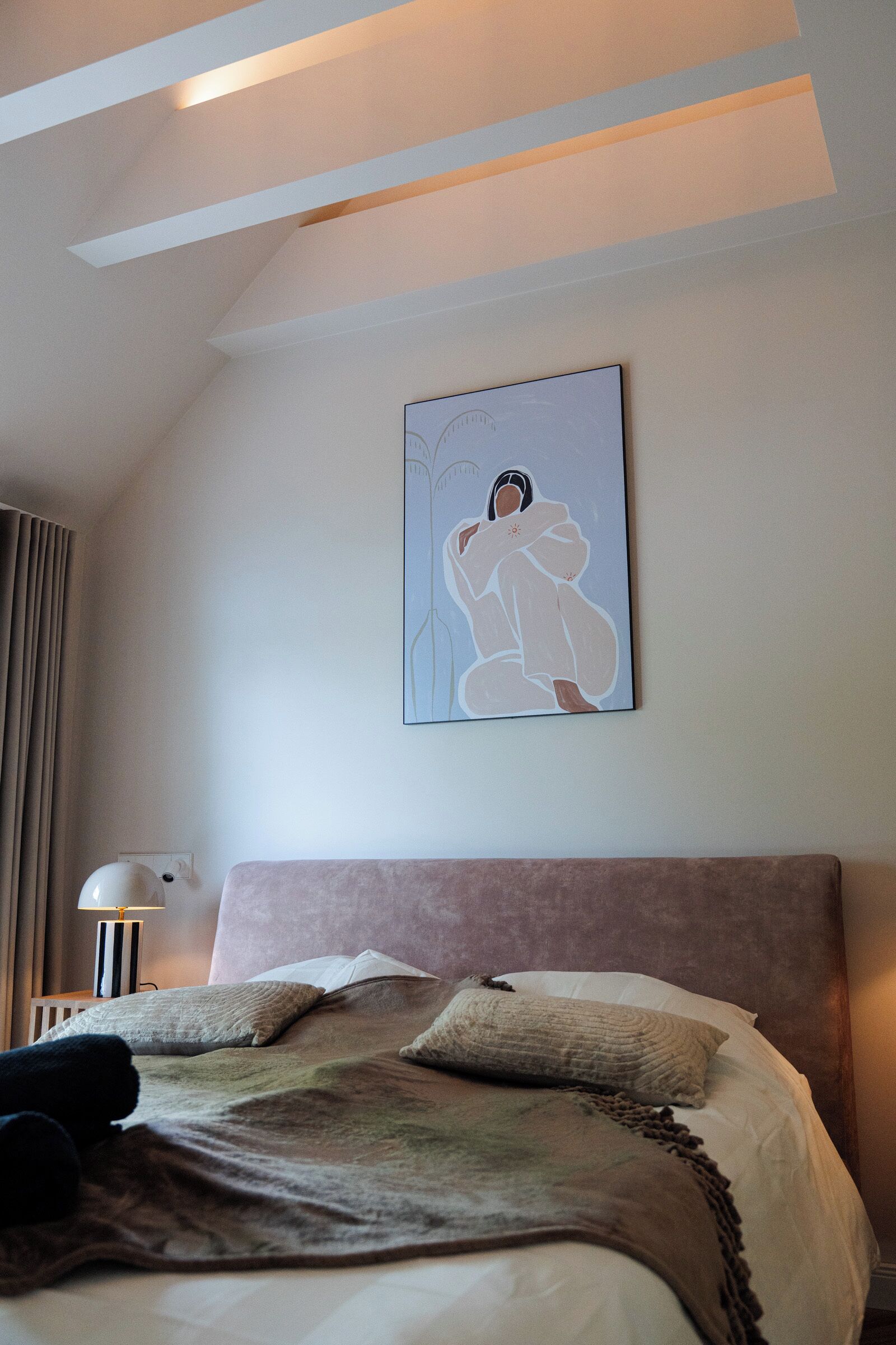 Ein Schlafzimmer mit Bett, Kissen, einem Nachttisch, einer Lampe und einem Wandbild.