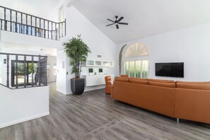 A spacious living area with a large sofa, a potted plant, a ceiling fan, and a TV mounted on the wall. - King Palm|Amazing Pool • Game Room • Near Kierland (Scottsdale)