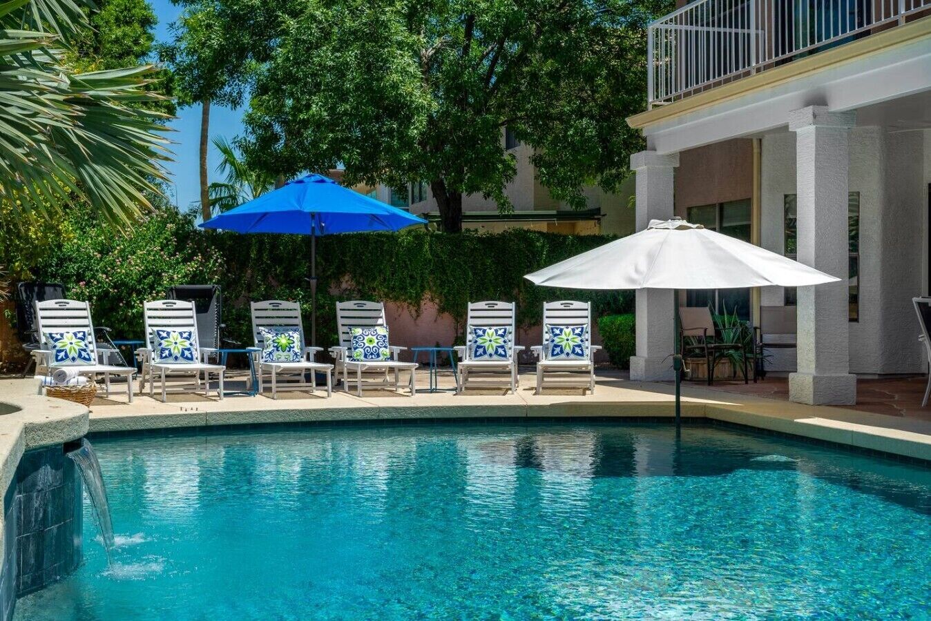 Un espace piscine doté de chaises longues et de parasols, entouré d’arbres et d’un bâtiment.