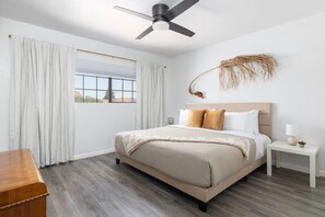 Een slaapkamer met een bed, gordijnen, een plafondventilator, een nachtkastje met een lamp en een decoratief object aan de muur.