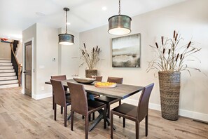 Un comedor con una mesa de madera, sillas, luces colgantes, un jarrón grande y un cuadro en la pared.