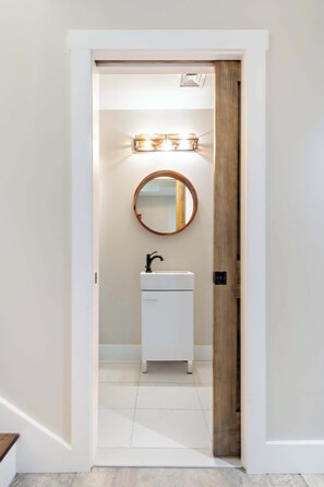 A hallway leading to a bathroom with a white sink, a round mirror, and a wooden door.