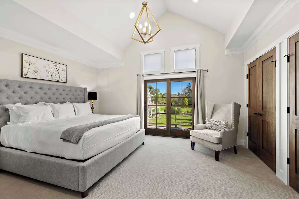 A bedroom with a large bed, a chair, a chandelier, and a view through a glass door.