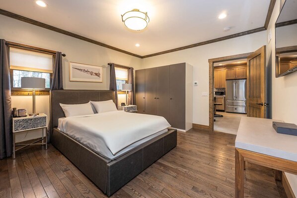 A modern bedroom with a large bed, bedside tables, a desk, and a view into the kitchen.