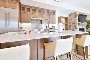 A modern kitchen with a central island, bar stools, a stone fireplace, and a TV mounted above it. - Hilltop | Village Two Bedroom Suite (Richmond)