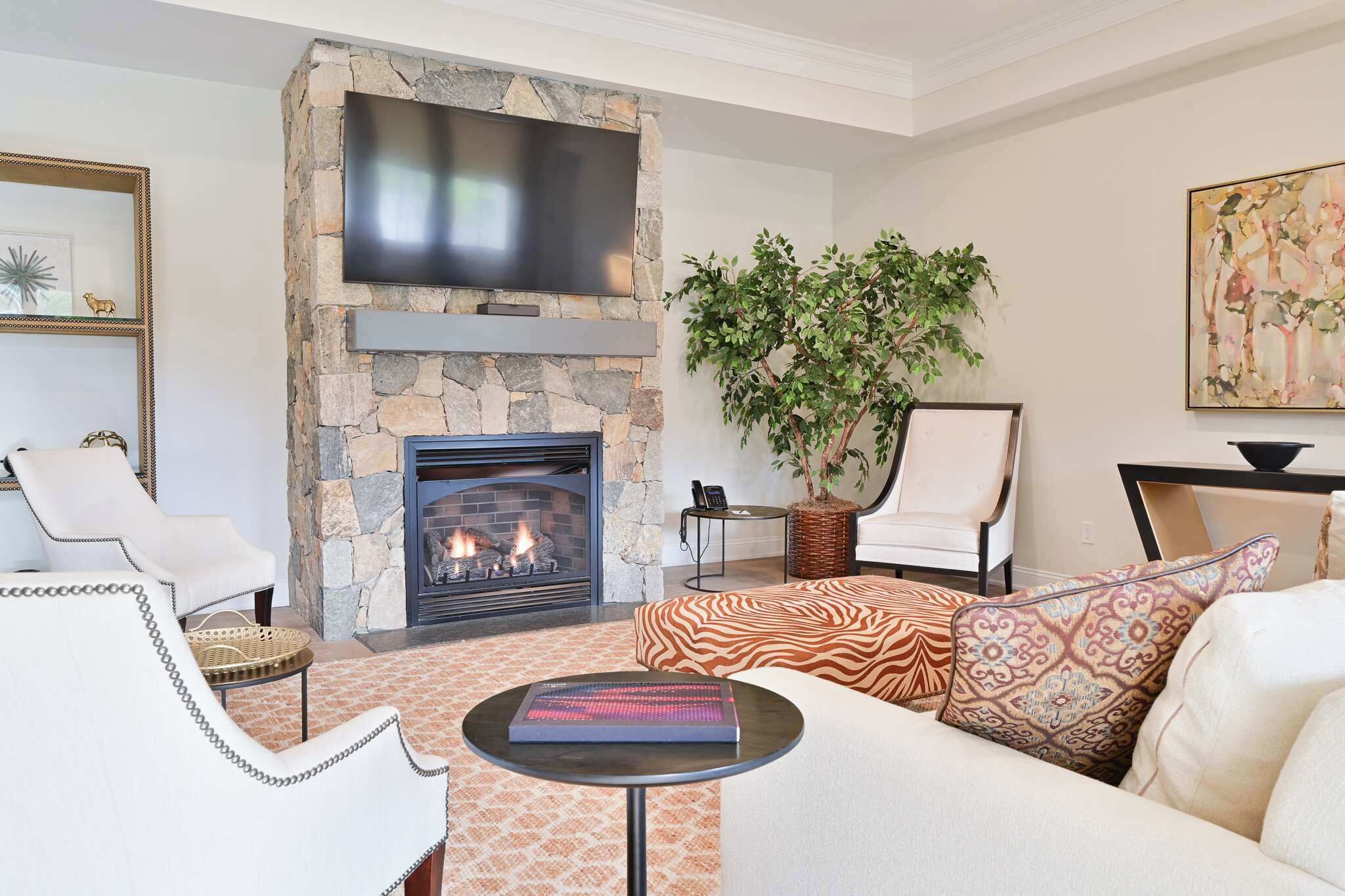 A living room with a stone fireplace, a flat-screen TV mounted above it, a comfortable sofa, a white armchair, a side table with a book, and a potted plant.