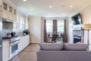 A modern kitchen with white cabinets, a dining area with a table and chairs, a gray sofa, and a stone fireplace. - Hilltop | Standard Queen Suite (Richmond)