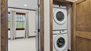 Une buanderie équipée de deux machines à laver et deux sèche-linge superposés, avec des portes en bois et une vue, par une porte ouverte, sur une pièce dotée d’une fenêtre et de rideaux.