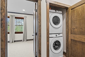 A laundry room with two stacked washers and dryers, wooden doors, and a view through an open door to a room with a window and curtains.