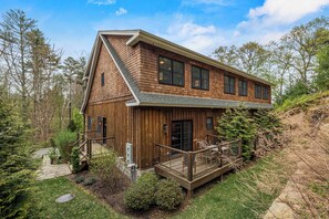A two-story wooden house with a deck, surrounded by trees and a stone pathway.