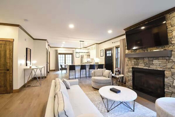 A spacious living room with a fireplace, a television mounted on a stone wall, and a dining area in the background.