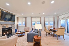 A spacious living room with a fireplace, a television mounted on a stone wall, and a dining area with a table and chairs. - Hilltop | Woodland Two Bedroom Suite (Richmond)