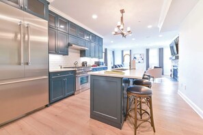 A modern kitchen with stainless steel appliances, dark blue cabinets, and a central island with bar stools. - Hilltop | Village Suite (Richmond)