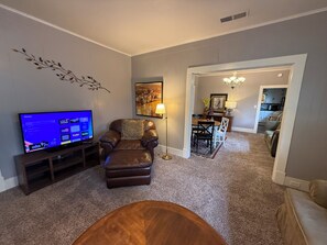 A living room with a flat-screen TV, leather armchair, and a dining area with a chandelier.