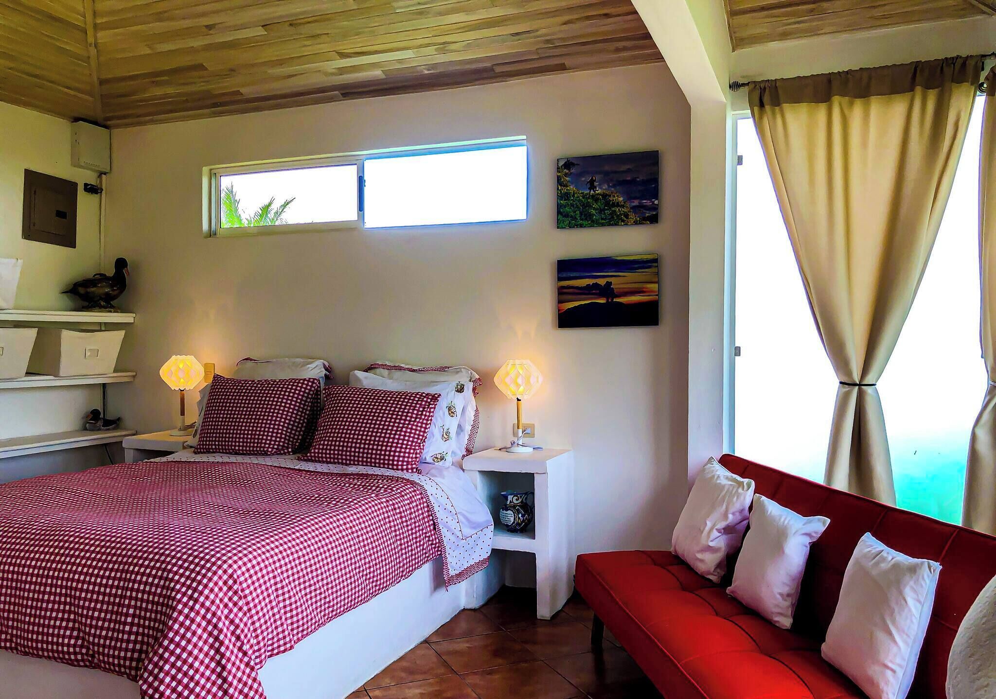 A bedroom with a bed, bedside tables, a red sofa, a window, and wall art.