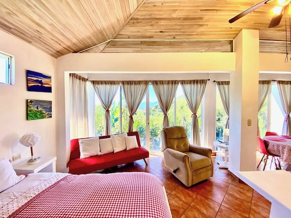 A room with a bed, a red sofa, a chair, and large windows with curtains.