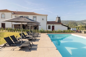 Un espace aménagé au bord de la piscine, avec des chaises longues et des parasols, une piscine et un bâtiment doté d’un toit en tuiles.