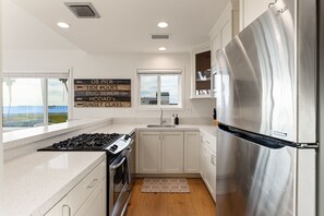 A modern kitchen with white cabinets, a stainless steel refrigerator, a gas stove, and a window with a view of the beach. - The Famous Big Blue Units 1 & 4 (San Diego)