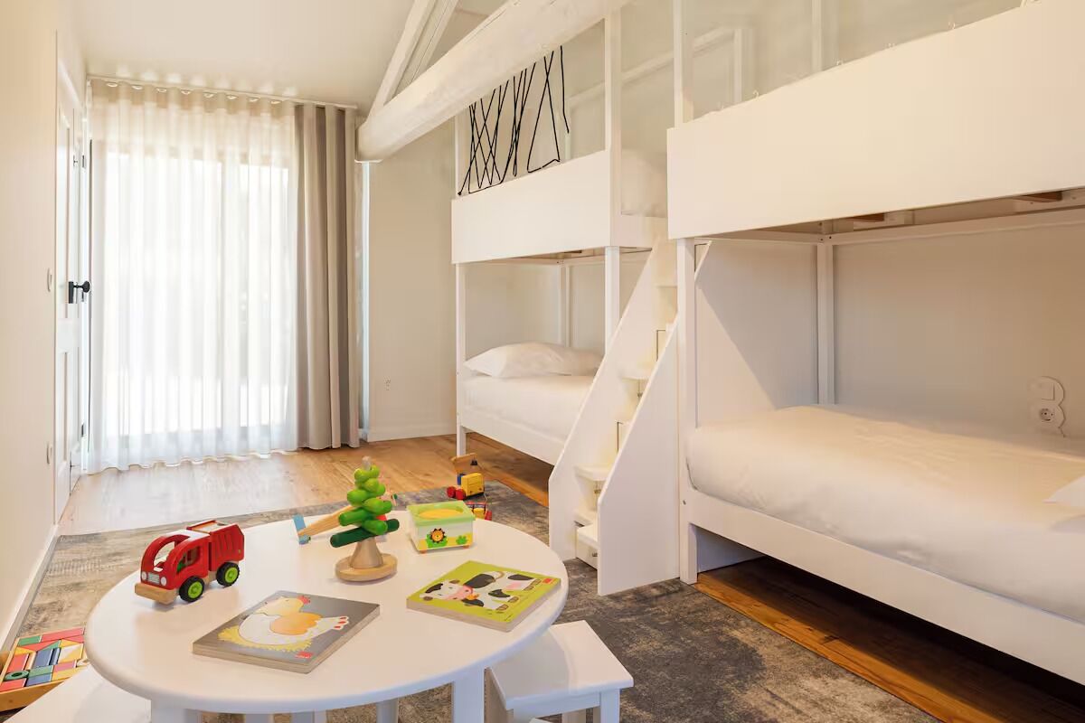A children's room with bunk beds, a small table with toys and books, and a window with sheer curtains.