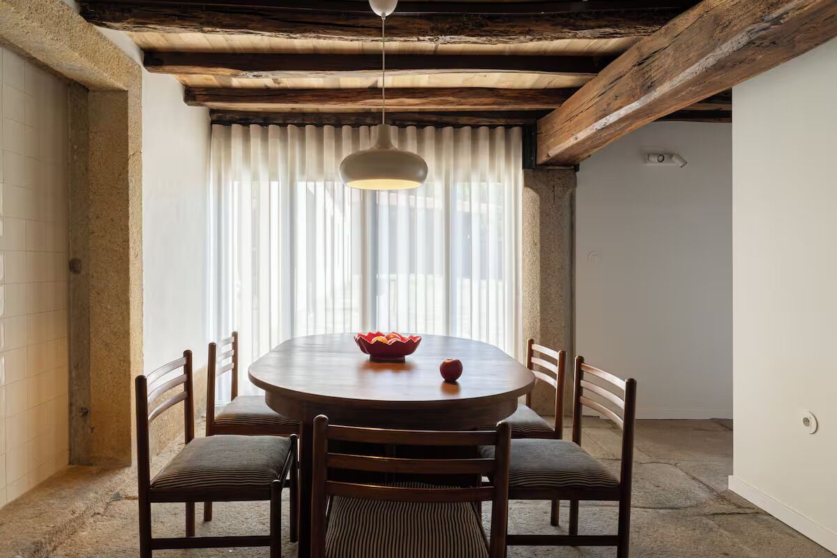 Ein Essbereich mit einem runden Holztisch, sechs Stühlen, einer hängenden Pendelleuchte und einem Fenster mit vertikalen Jalousien.
