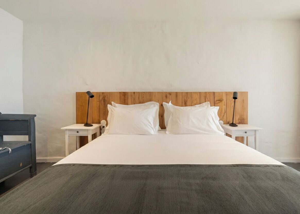 Een slaapkamer met een houten hoofdbord, twee nachtkastjes met lampen, een bed met witte lakens en een donker kastje.