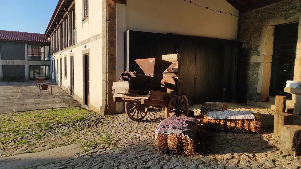 A rustic building with a stone facade, a wooden cart, hay bales, and a covered outdoor area.