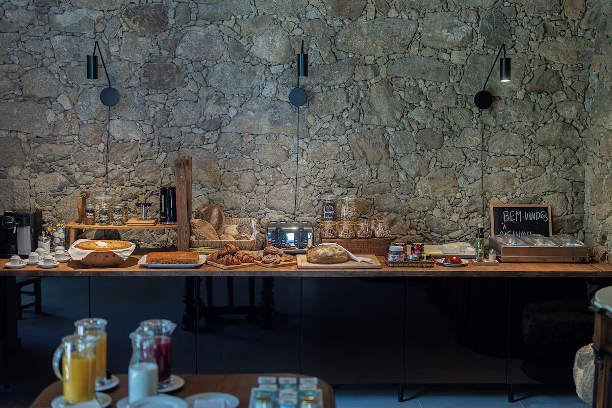 A breakfast buffet with a stone wall backdrop, a coffee station, and a sign reading "BEM-VINDOS A CASA NOSTRA".