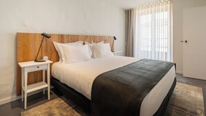 A modern bedroom with a wooden headboard, a white bedspread, a black accent pillow, a bedside table with a lamp, and a large window with sheer curtains. - Rustic Countryside Stay with Kitchen, Living & Dining Area, + Swimming Pool (Braga)