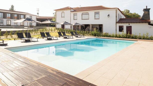 A swimming pool with lounge chairs and umbrellas, surrounded by a wooden deck and a white building with a tiled roof. - Rustic Countryside Stay with Kitchen, Living & Dining Area, + Swimming Pool (Braga)