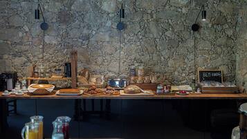 A breakfast buffet with a stone wall backdrop, a coffee station, and a sign reading "BEM-VINDOS A CASA NOSTRA".