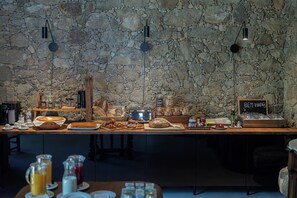 A breakfast buffet with a stone wall backdrop, a coffee station, and a sign reading "BEM-VINDOS A CASA NOSTRA".
