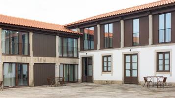 A two-story building with large windows and a tiled roof. There are two doors and a small outdoor seating area with chairs.