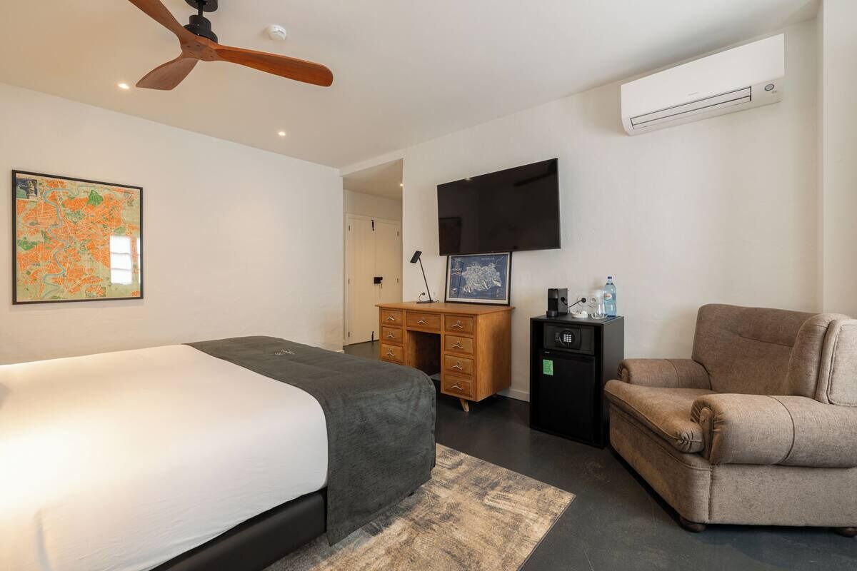 A modern hotel room with a bed, a desk, a chair, a flat-screen TV, and an air conditioning unit.