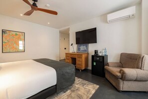 A modern hotel room with a bed, a desk, a chair, a flat-screen TV, and an air conditioning unit.